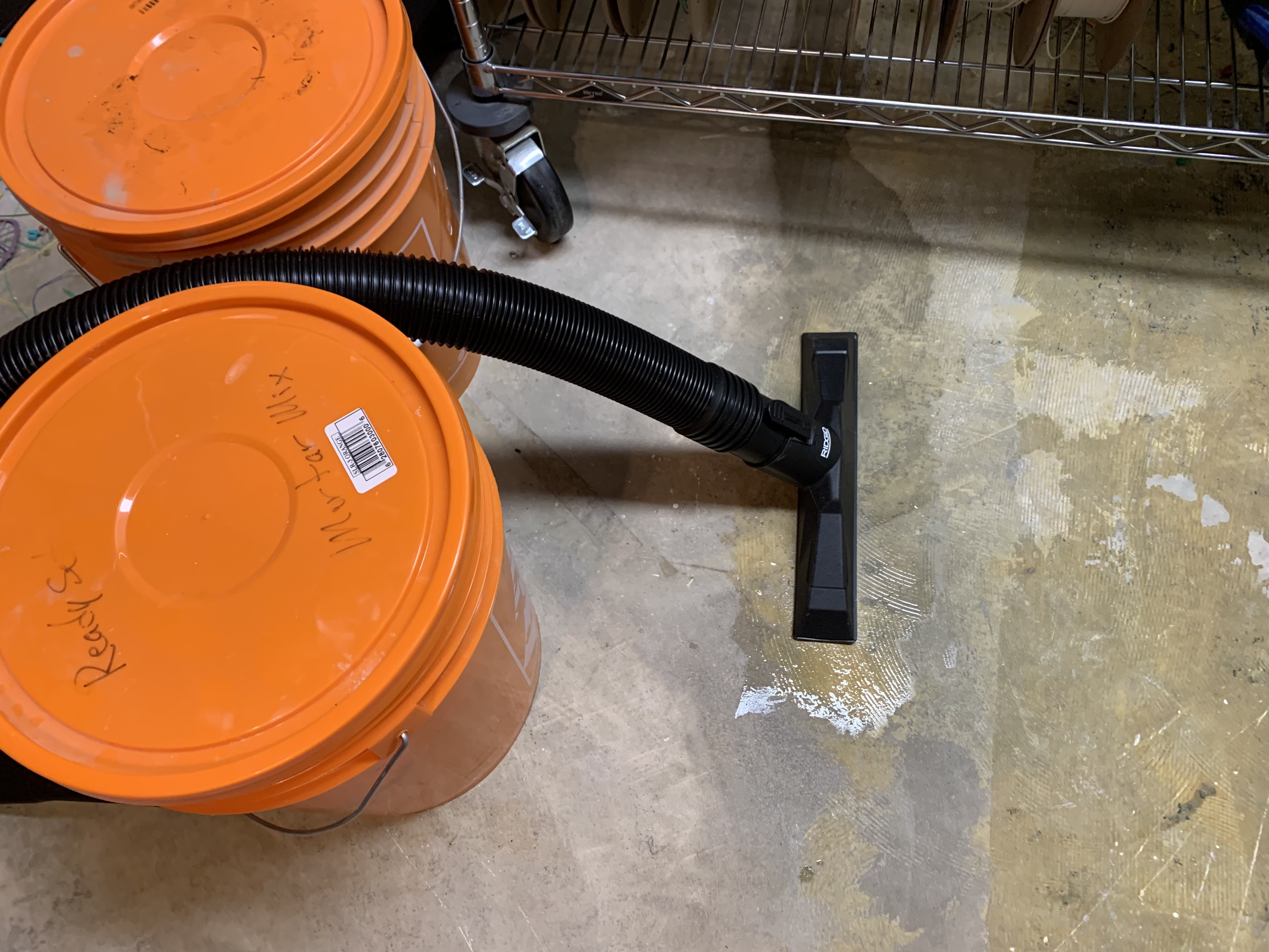 A shop vac floor nozzle held in place over a thin stream of water on a concrete workshop floor, pinned by two orange 20-litre cement pails labelled 'Mortar Mix' and 'Ready S'.